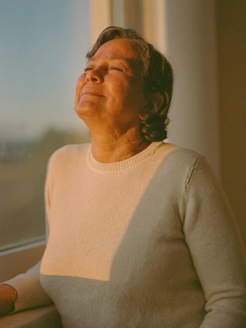 Una mujer junto a la ventana con los ojos cerrados, iluminada por el sol de la mañana